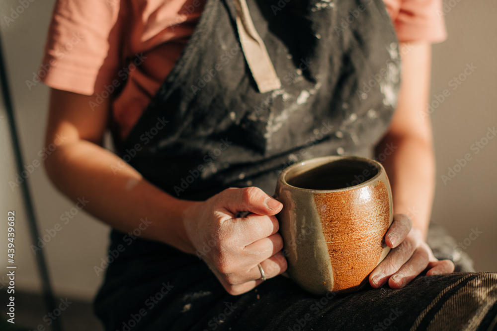 Female potter in apron caked with clay holding a cup of tea Female potter in apron caked with clay holding a cup of tea
