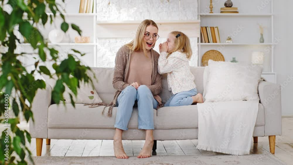 Adult mother sits on sofa at home in living room with little girl daughter covering mouth with hands talking whispering secret to share secrecy information mommy says wow surprised, trust concept Adult mother sits on sofa at home in living room with little girl daughter covering mouth with hands talking whispering secret to share secrecy information mommy says wow surprised, trust concept