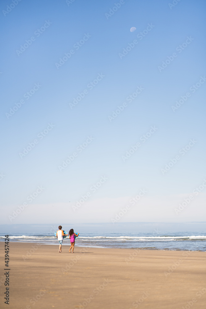 Siblings walk towards the ocean carrying skimboards Siblings walk towards the ocean carrying skimboards