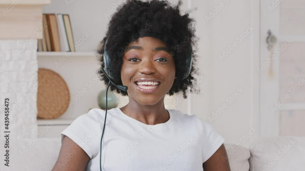 Smiling African young woman wears headset talks to web camera making distance online video conference call. Female internet teacher doing distant chat working from home. Telework concept. Webcam view Smiling African young woman wears headset talks to web camera making distance online video conference call. Female internet teacher doing distant chat working from home. Telework concept. Webcam view