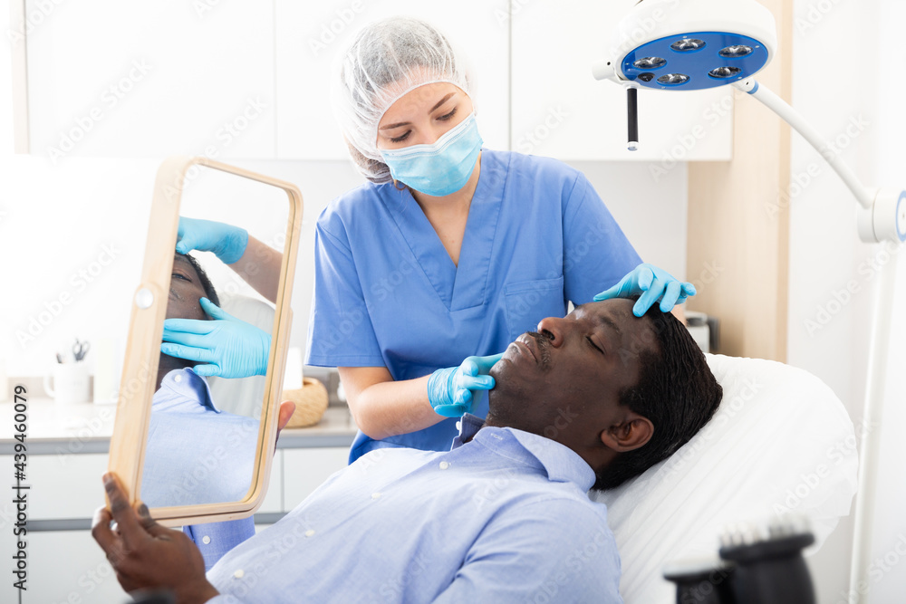 Fototapeta premium Skilled female beautician in face mask giving consultation to african male patient in aesthetic clinic