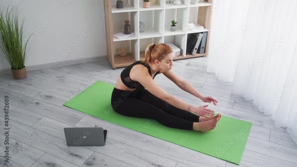 Pilates online. Training at home. Healthy lifestyle. Athletic woman in sportswear doing stretching fitness exercise with laptop on yoga mat on floor at light modern room interior. Pilates online. Training at home. Healthy lifestyle. Athletic woman in sportswear doing stretching fitness exercise with laptop on yoga mat on floor at light modern room interior.