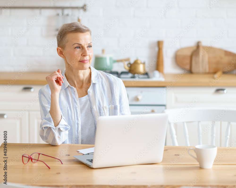 Happy dreaming senior woman in casual clothes looking aside and thinking while working or surfing internet on laptop, happy elderly 60s woman enjoying morning with coffee in light modern kitchen Happy dreaming senior woman in casual clothes looking aside and thinking while working or surfing internet on laptop, happy elderly 60s woman enjoying morning with coffee in light modern kitchen