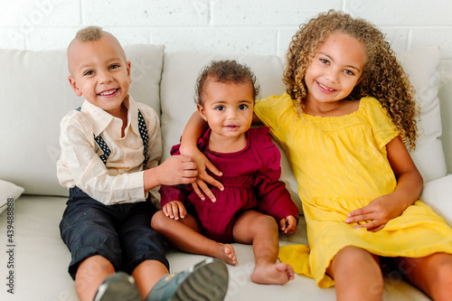 Three young smiling biracial siblings sitting together on white couch