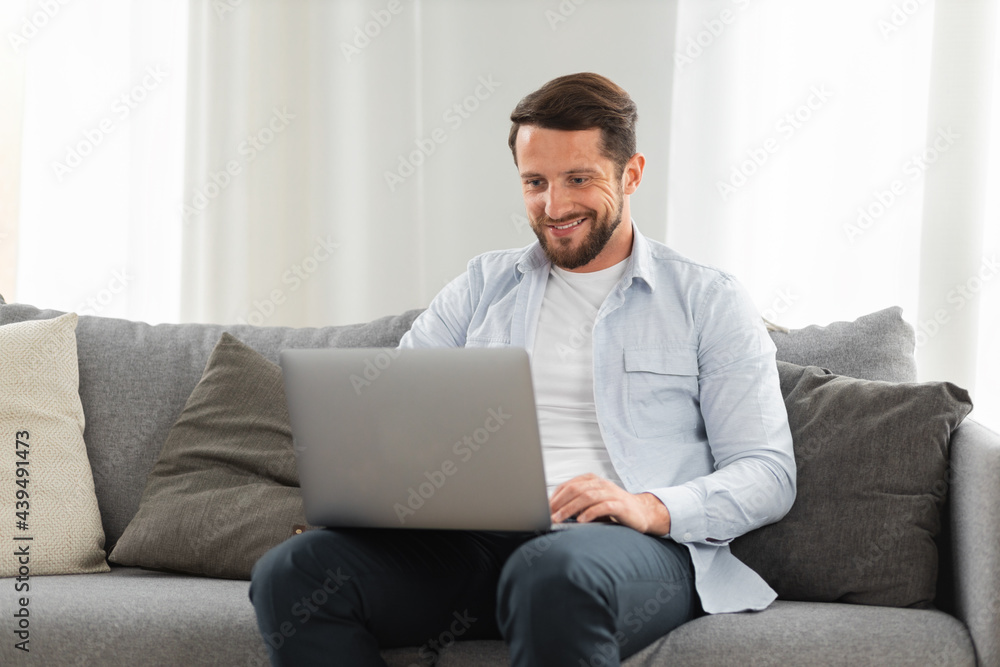 Happy caucasian man uses laptop computer for video call while sitting on sofa at home, smiling caucasian man chatting with friends or family via video conference on social networks Happy caucasian man uses laptop computer for video call while sitting on sofa at home, smiling caucasian man chatting with friends or family via video conference on social networks