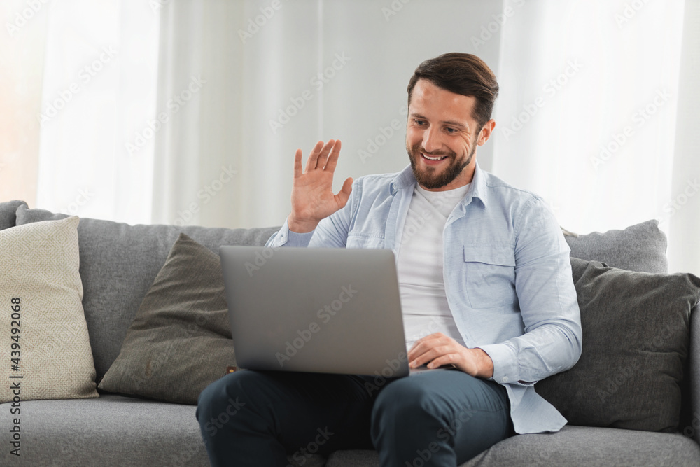 Happy caucasian man uses laptop computer for video call while sitting on sofa at home, smiling caucasian man chatting with friends or family via video conference on social networks, greeting gesture Happy caucasian man uses laptop computer for video call while sitting on sofa at home, smiling caucasian man chatting with friends or family via video conference on social networks, greeting gesture