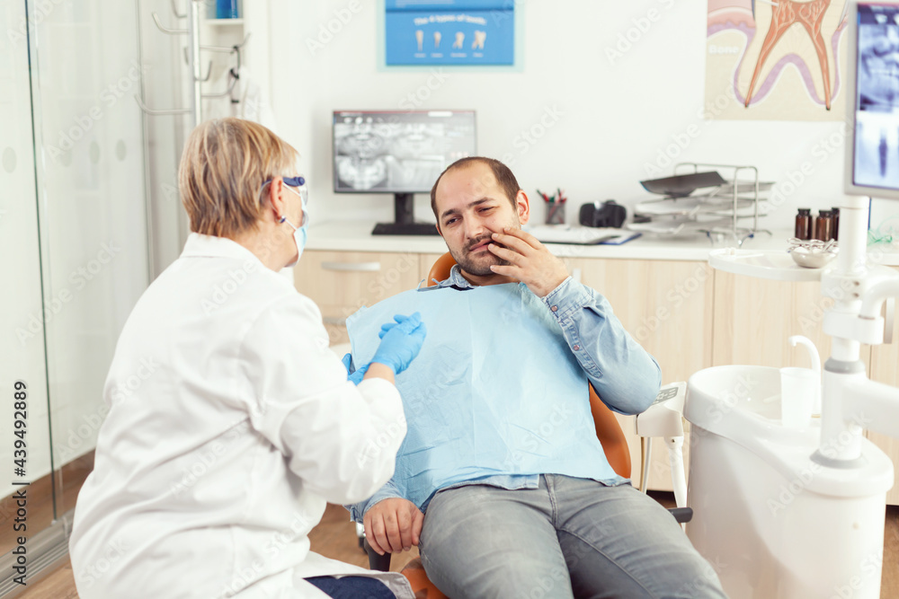 Sick man complaining about tooth pain discussing with stomatologist senior woman about stomatology treatment. Orthodontic doctor preparing for dentistry surgery during medical appointment Sick man complaining about tooth pain discussing with stomatologist senior woman about stomatology treatment. Orthodontic doctor preparing for dentistry surgery during medical appointment