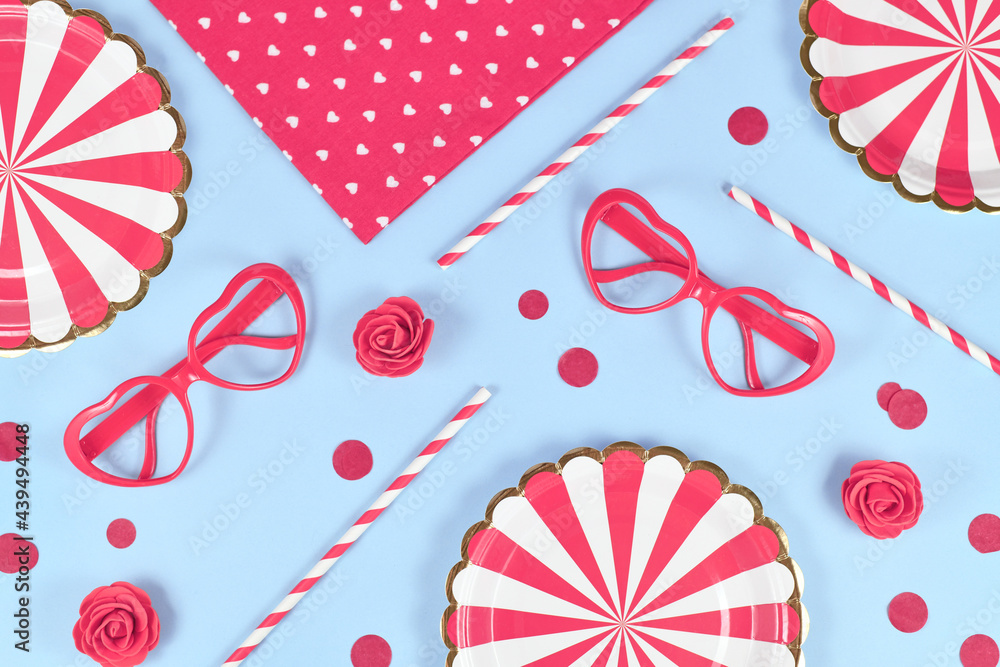Obraz premium Party flat lay with paper plates, heart shaped glasses, roses, confetti and drinking straws on blue background