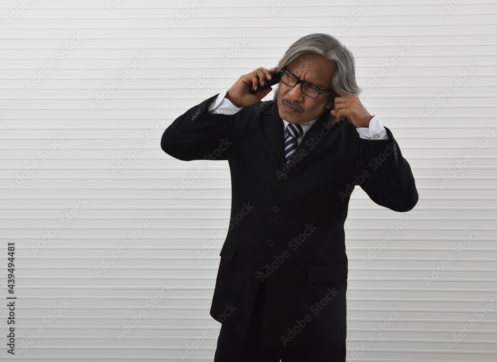 Thoughtful asian senior businessman in black suit and eyeglasses holding smart mobile phone and touching his face with finger standing over white wall background, Business communication concept Thoughtful asian senior businessman in black suit and eyeglasses holding smart mobile phone and touching his face with finger standing over white wall background, Business communication concept