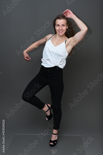 Portrait of a Caucasian girl with fluttering hair in the wind in black trousers and a white blouse with a smile on a gray background in full growth. Studio photo in high resolution.