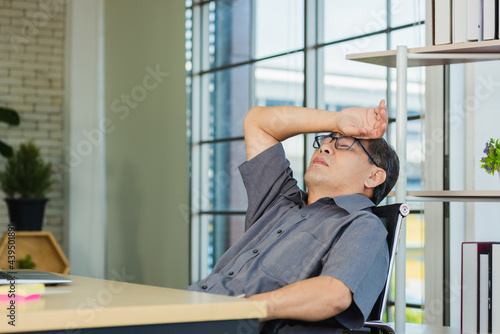 Asian businessman tired overworked he relaxing with closed eyes and hands behind his head on the desk. senior man with eyeglasses lying asleep on table at his working place photo