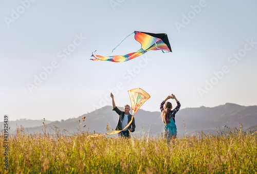 Daughter with smiling father while they flying a colorful kites on the high grass meadow in the mountain fields. Warm family moments or outdoor time spending concept image. photo