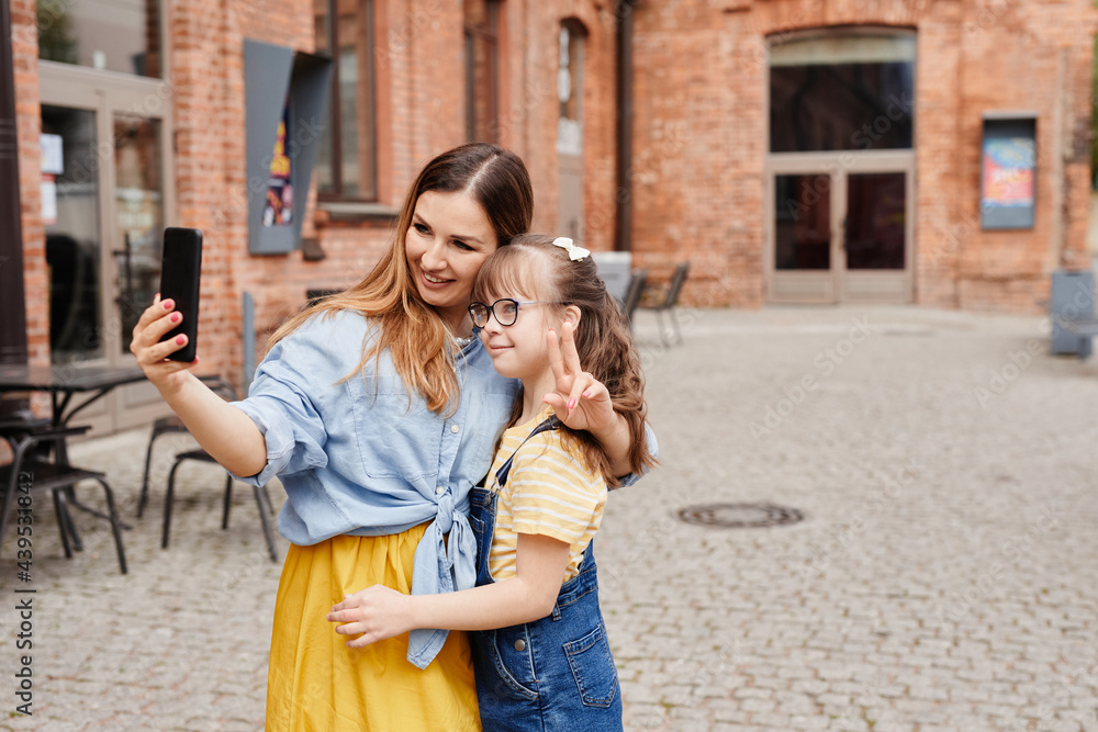 Fototapeta premium Colorful waist up portrait of happy mother and daughter with down syndrome taking selfie together outdoors in city, copy space