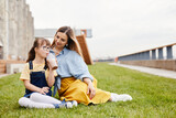 Full length portrait of happy mother and daughter with down syndrome sitting on green lawn outdoors together, copy space