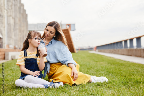 Full length portrait of happy mother and daughter with down syndrome sitting on green lawn outdoors together, copy space