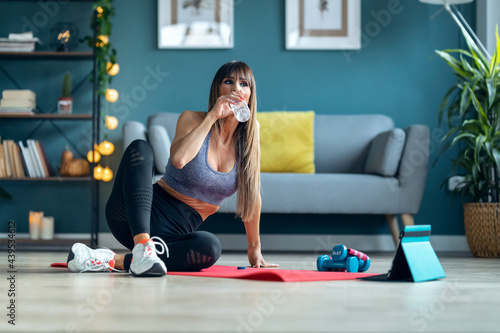 Sporty mature woman drinking water while making a break after online gym classes in the living room at home.