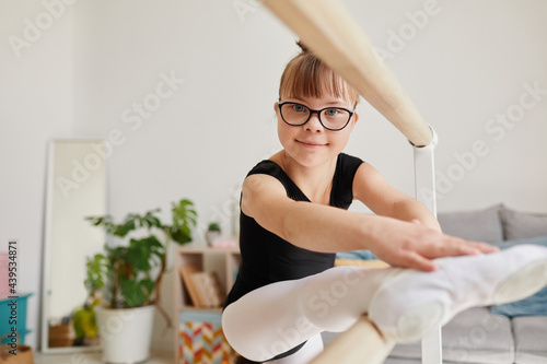 Portrait of cute ballerina with down syndrome standing by bar at home and doing stretching exercise, copy space