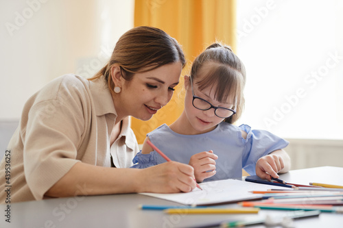 Fototapeta Portrait of cute girl with down syndrome studying at home with caring mother hel