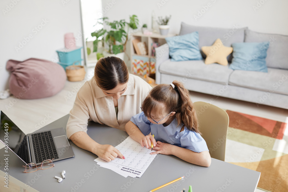 High angle portrait of cute teenage girl with down syndrome doing homework at desk with caring mother helping her High angle portrait of cute teenage girl with down syndrome doing homework at desk with caring mother helping her