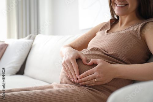 Pregnant young woman making heart with her hands on belly at home, closeup