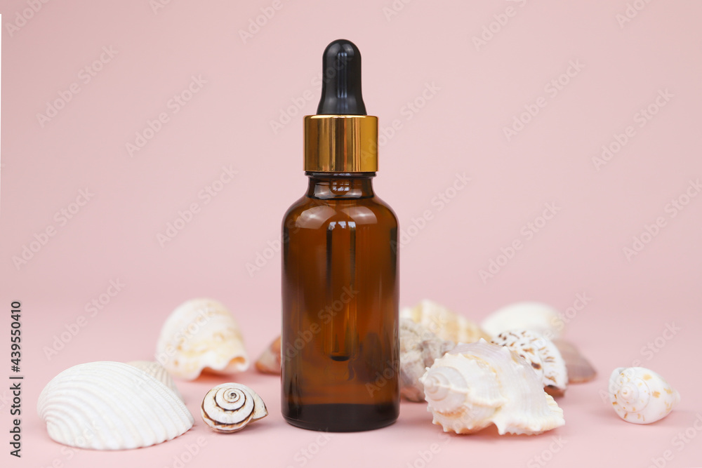 Dropping essential oil into glass bottle or bottle of hyaluronic acid with pipette, many different sea shells on pink background. Serum for facial skin. Unbranded herbal mineral cosmetic. Front view. Dropping essential oil into glass bottle or bottle of hyaluronic acid with pipette, many different sea shells on pink background. Serum for facial skin. Unbranded herbal mineral cosmetic. Front view.