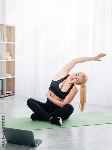 Stretching exercise. Female home sport. Online training. Happy athletic woman in black sportswear lotus pose doing warming workout side bending looking laptop in light room interior.