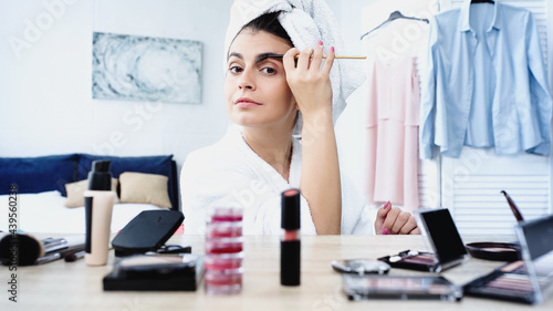 young woman in bathrobe with head wrapped in towel styling eyebrow with brush near table with decorative cosmetics in bedroom