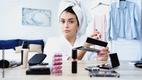 young woman in bathrobe sitting with cosmetic brush and eye shadows near table with decorative cosmetics in bedroom