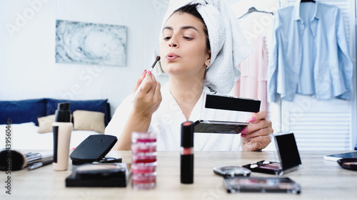 young woman in bathrobe blowing on cosmetic brush near table with decorative cosmetics in bedroom © LIGHTFIELD STUDIOS