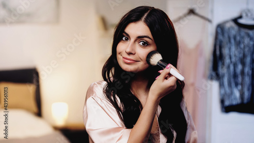smiling young woman in pink silk peignoir applying face powder with cosmetic brush in bedroom