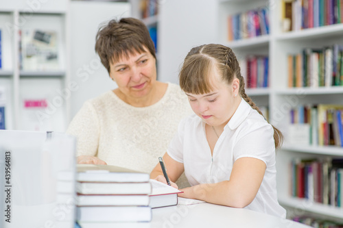 Senior woman helps to girl with syndrome doing homework at library. Education for disabled children concept
