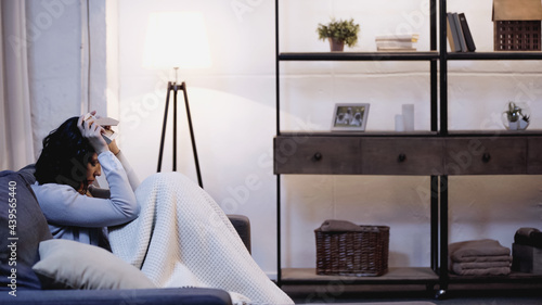 depressed woman covered in blanket sitting on sofa and holding photo frame near head at home