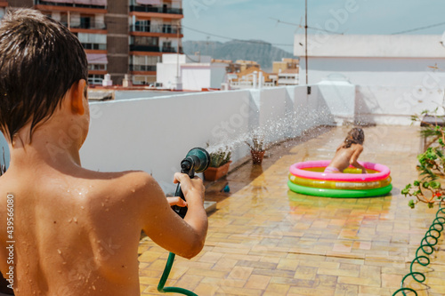 funny boy with hose refreshing with hosepipe to sister in terrace into a inflatable pool