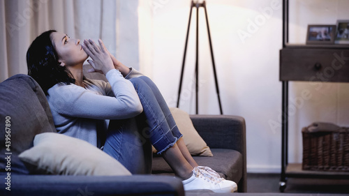 Bild auf Leinwand depressed woman in casual clothes sitting on sofa with praying hands near mouth