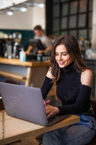 Young female with cute smile sitting with portable net-book in modern coffee shop interior during recreation time, charming happy woman student using laptop to prepare for the course work
