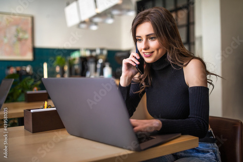 Happy young woman talking by mobile phone and using laptop at cafe