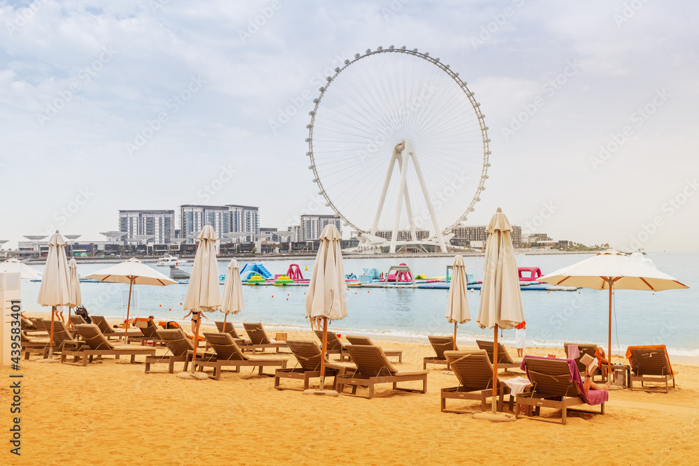 Golden sand between rows of sun chairs and beach umbrellas in the JBR district in Dubai. Ferris wheel at the background. Recreation and vacation in the UAE Golden sand between rows of sun chairs and beach umbrellas in the JBR district in Dubai. Ferris wheel at the background. Recreation and vacation in the UAE