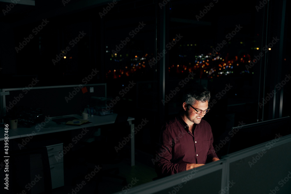 Middle age businessman working on a computer while sitting in a dark office at night with city lights in the background Middle age businessman working on a computer while sitting in a dark office at night with city lights in the background
