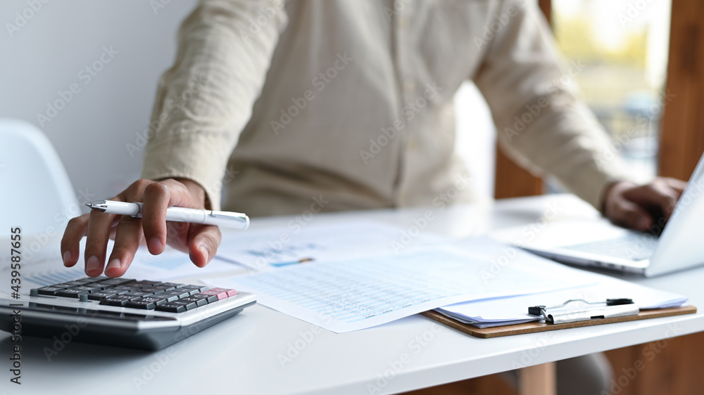 Hand with pen to use calculator and laptop keyboard, graphs and file folders on the desk. Hand with pen to use calculator and laptop keyboard, graphs and file folders on the desk.