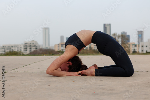 Pretty and flexible young woman bending her back in a Yoga pose. Shot in an empty urban park with Tel Aviv buildings in the far background.