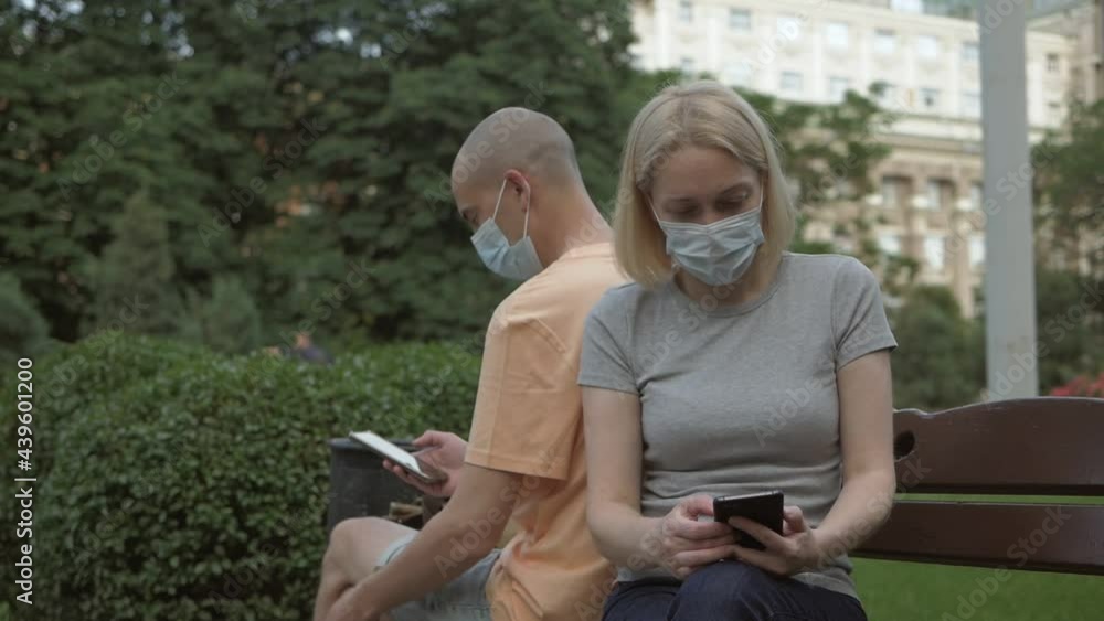 A man and a woman wearing protective masks sit on a bench and use their phones in a park A man and a woman wearing protective masks sit on a bench and use their phones in a park