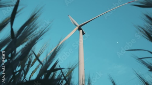 Windmill with rotating propeller produces clean electricity against blue sky on summer day view from green wheat low angle shot