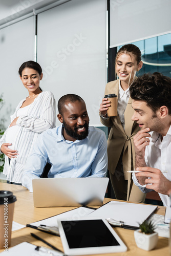 african american businessman smiling near laptop and positive colleagues during meeting