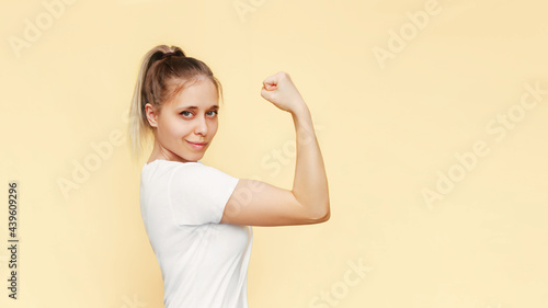 Strong powerful confident caucasian young blonde woman raises arm and shows bicep isolated on a light color beige background. Feminism, girl power, equal women's rights and independence concept