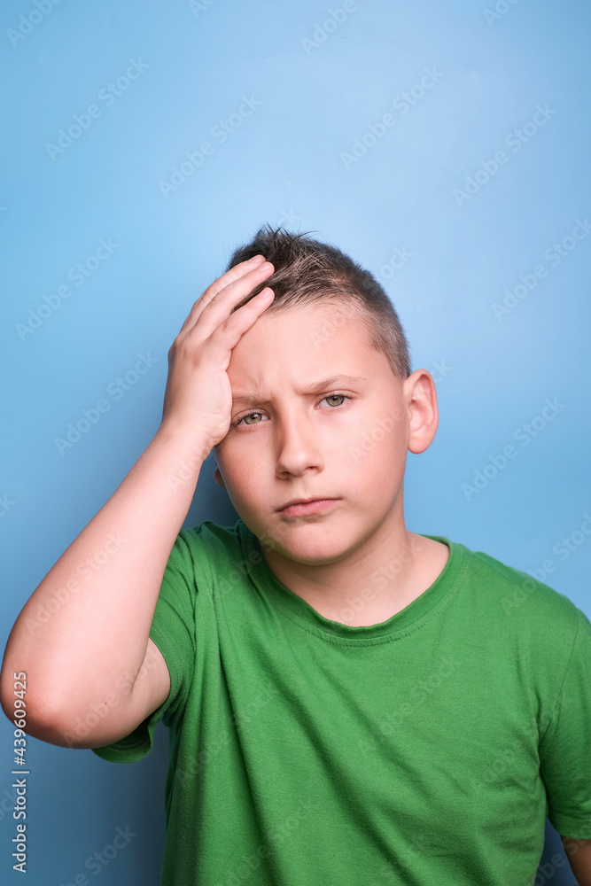 Studio shot of emotional adorable boy covering head with hand being tired, headache, showing true emotion. Portrait on blue background Studio shot of emotional adorable boy covering head with hand being tired, headache, showing true emotion. Portrait on blue background