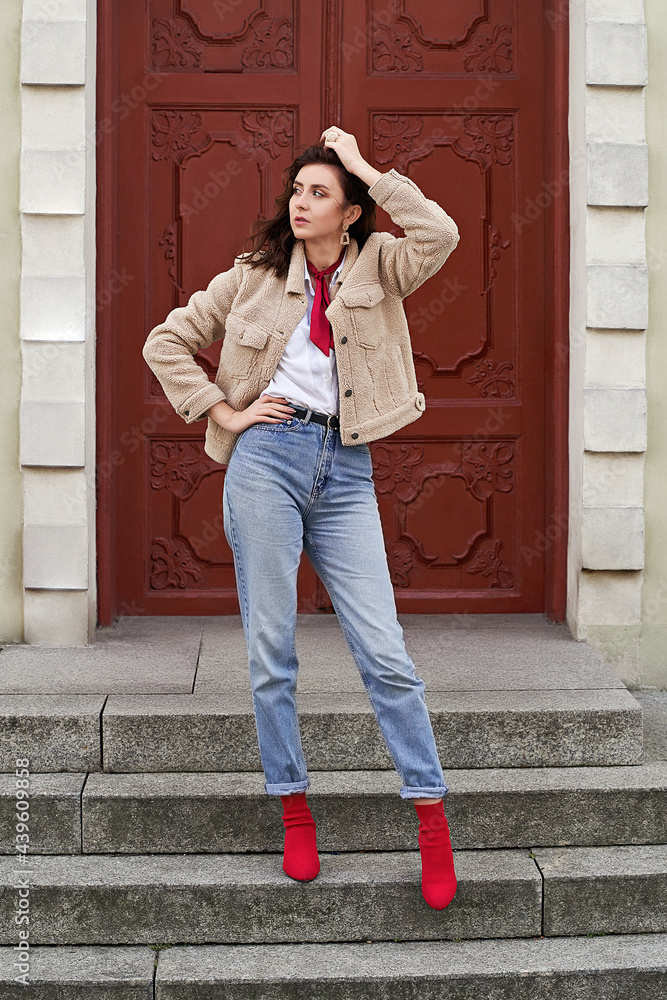 Young beautiful woman in red high heel shoes, blue denim jeans pants and teddy jacket coat posing on old red door background in European city