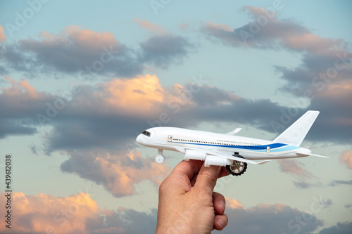 A hand holding a toy white airplane against the backdrop of a beautiful sky with clouds illuminated by the setting sun