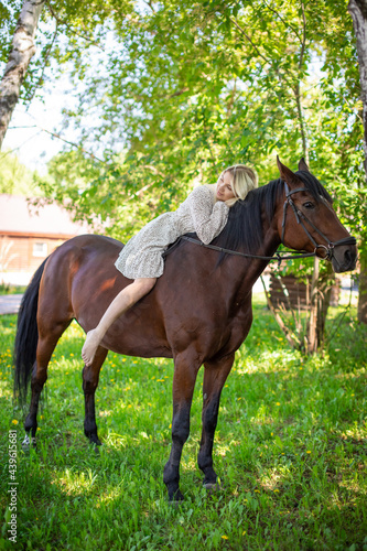 A young rider woman blonde with long hair in a dress posing with brown horse in forest next to farm