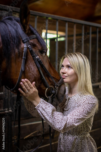 A young rider woman blonde with long hair in a dress posing with brown horse inside stable, Russia