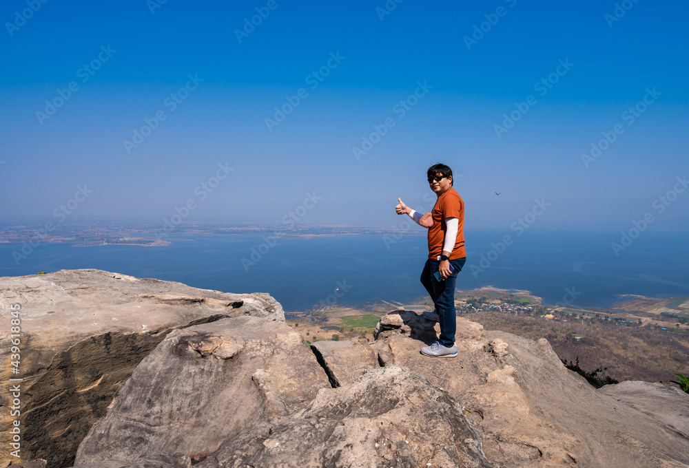 happy Asian man standing with happy smile and thumb up on top viewpoint on rock mountain peak with misty lake dam view and blue sky at Pha Muen viewpoint, Khon Kaen, Thailand, satisfy travel  concept